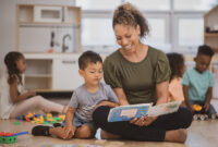 A preschool boy of Hispanic ethnicity sits next to his teacher as she is reading him a book on the classroom floor.