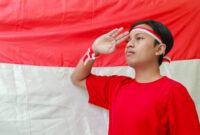 Portrait of attractive Asian man in red shirt with red and white ribbon on head, giving salute pose with hand in front of Indonesian flag